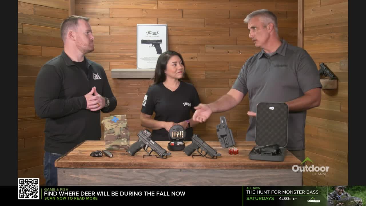 Three people stand behind a table displaying firearms and accessories. The man on the right gestures toward an open case, while the woman and the other man look on.
