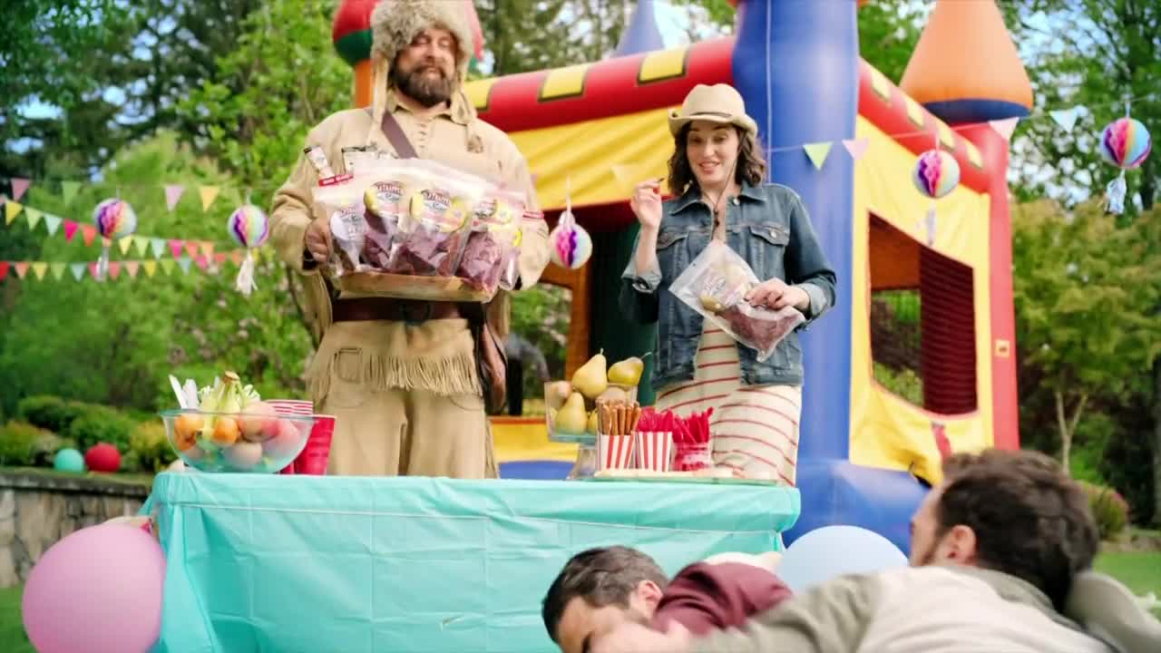 A man in a coonskin cap holds a tray of packaged meat, while a woman in a denim jacket gestures with a similar bag.  A bounce house and a table of snacks suggest a backyard party.
