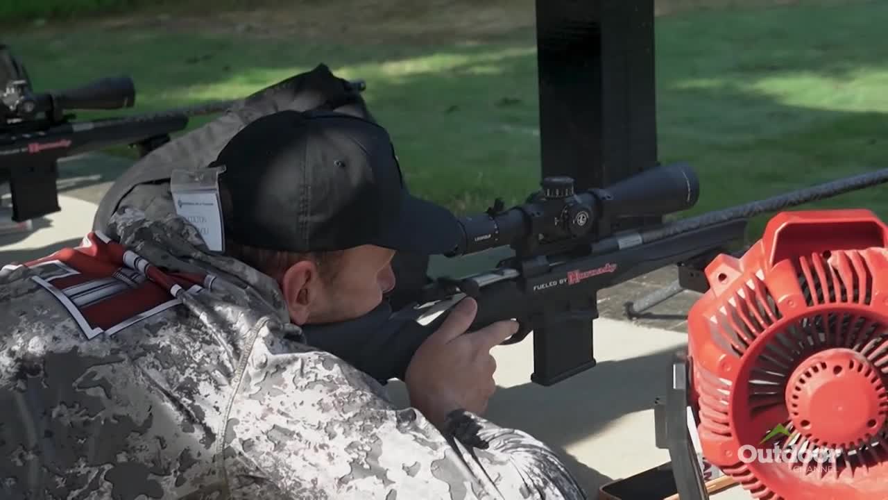 A man in camo gear is aiming a rifle, his eye pressed against the scope. The Outdoor Channel logo is visible, suggesting a competition or demonstration in the United States.
