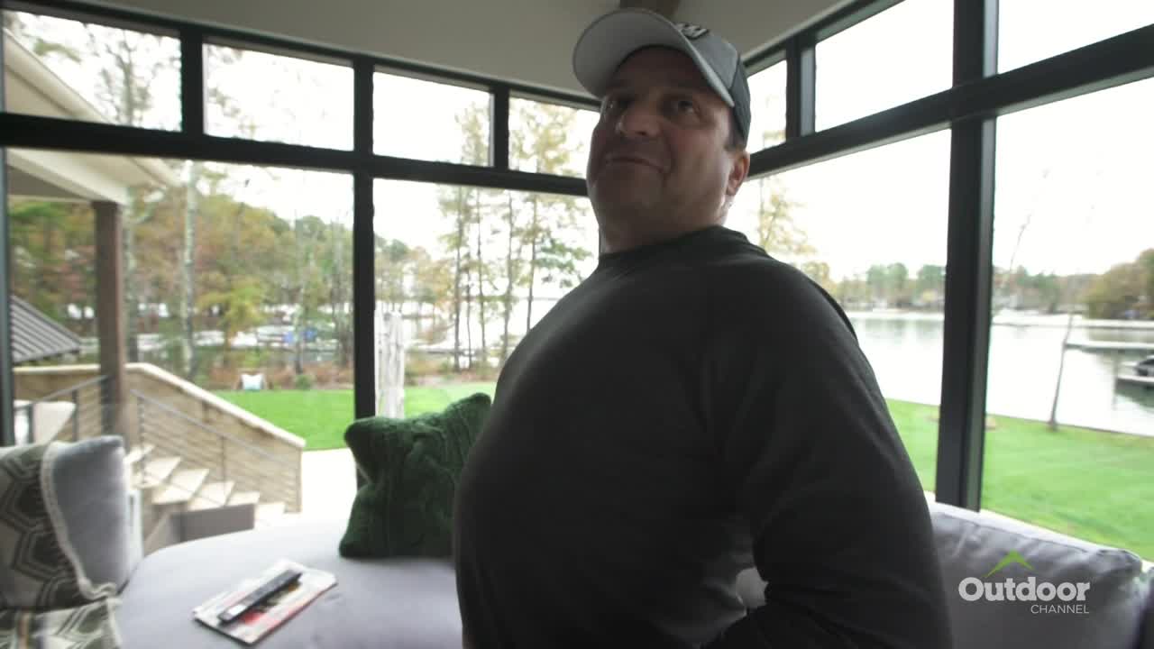 A man in a baseball cap and black shirt stands in a modern home, looking towards the camera. Through the large windows, a lake scene is visible, with the Outdoor Channel logo in the corner.
