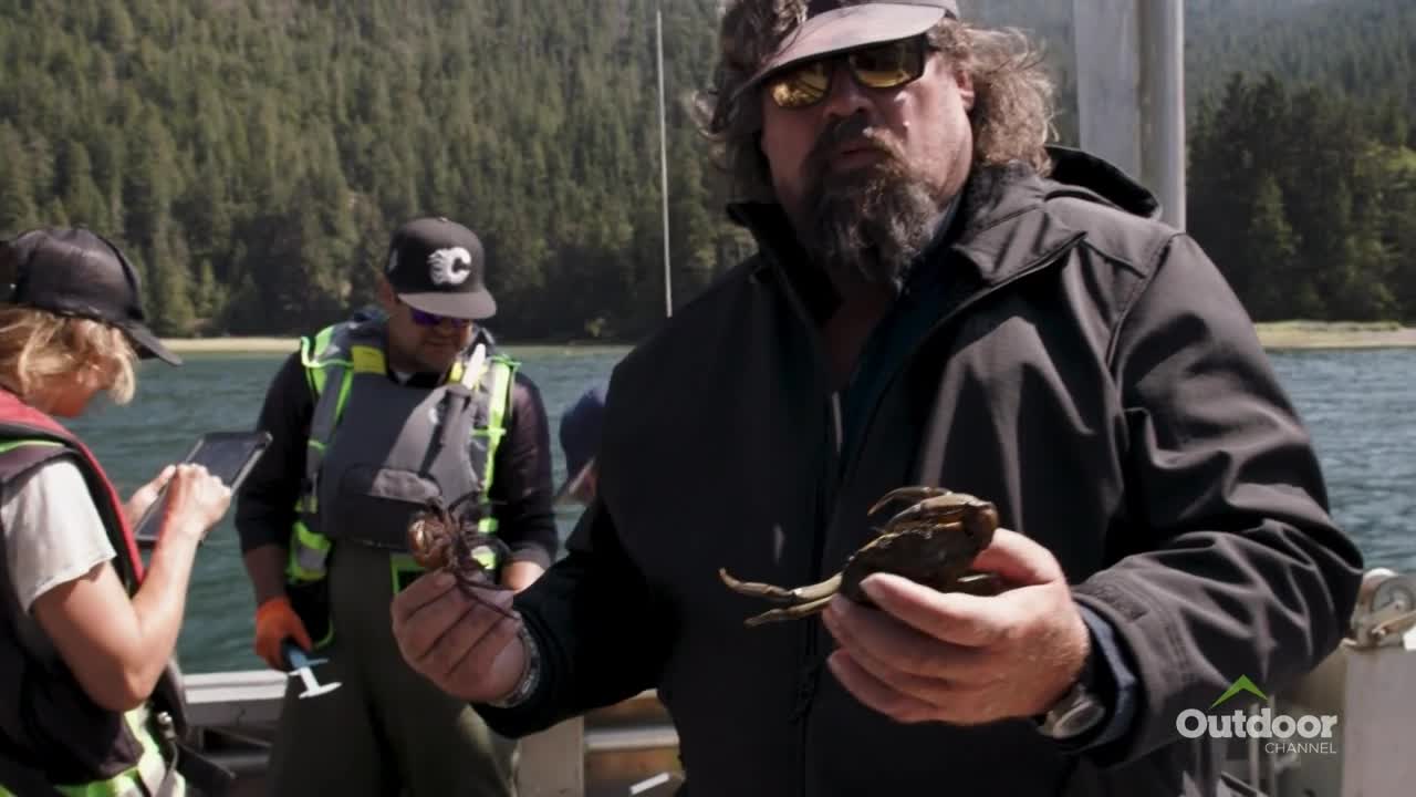 A man in sunglasses holds a crab up for the camera, while others on the boat look on. The Outdoor Channel logo is visible in the corner.
