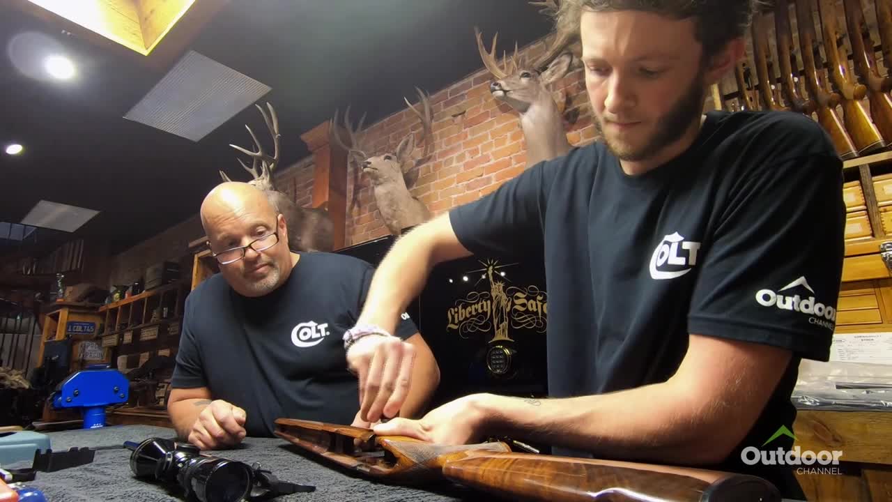 Two men, both wearing Colt shirts, are working on a rifle. One is carefully tightening something on the wooden stock while the other watches intently.
