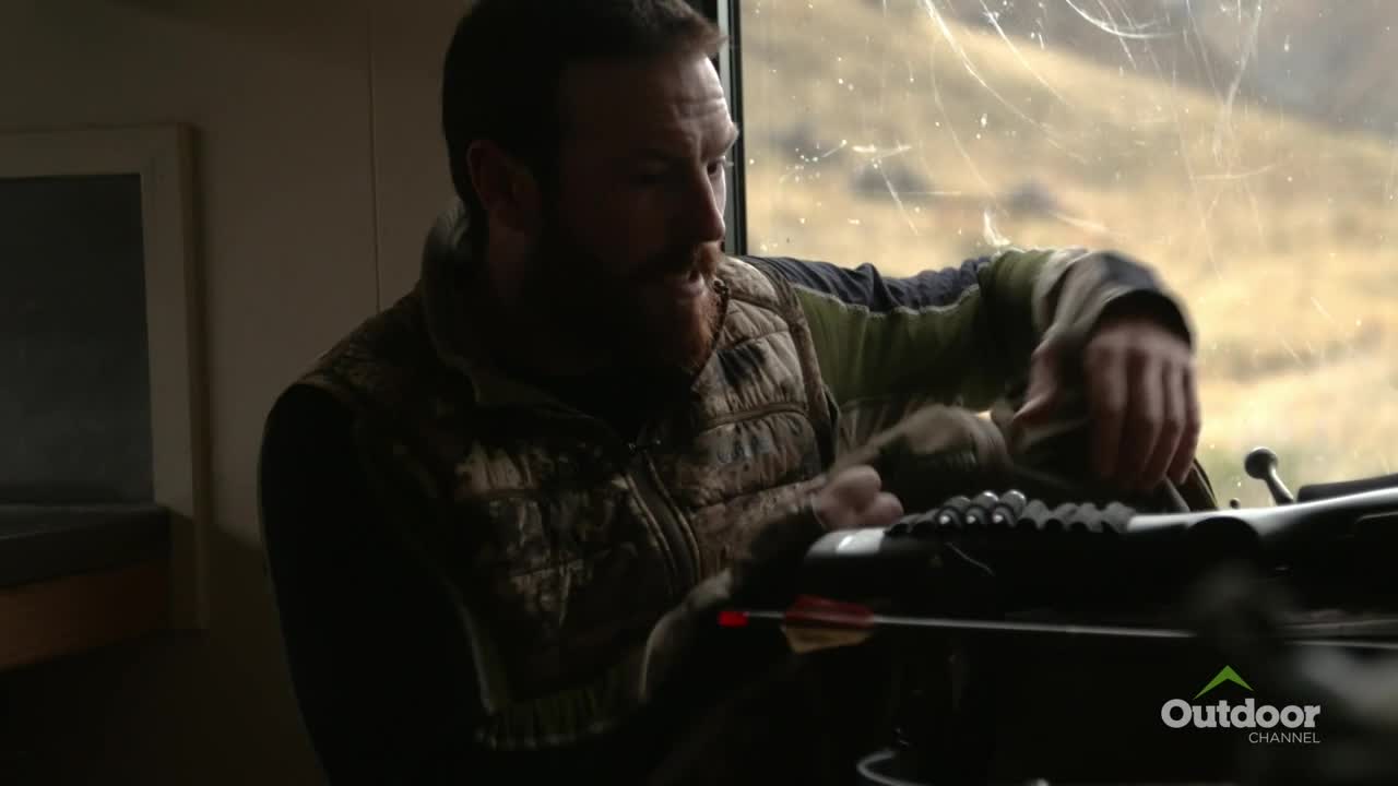 Inside a hunting blind, a man in a camouflage vest carefully handles a crossbow. An arrow is already loaded, and he seems focused on the task at hand.
