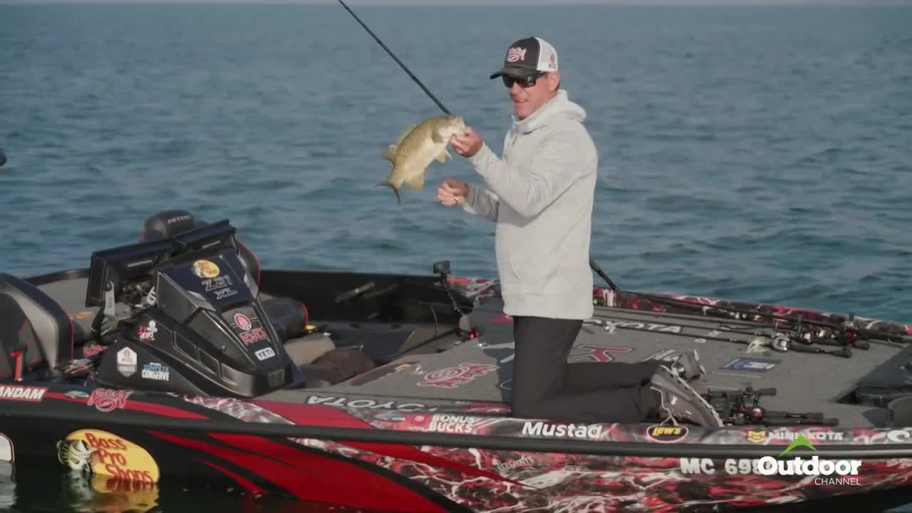 A man in a ball cap and sunglasses holds up a fish, its body arched. He's kneeling in a bass boat, the Outdoor Channel logo visible in the corner.
