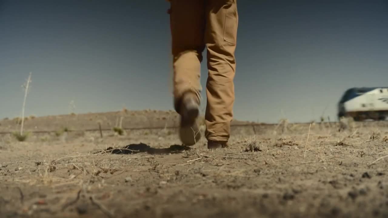 A person in work boots and tan pants strides across the dry, cracked earth. In the distance, a train speeds along the tracks.

