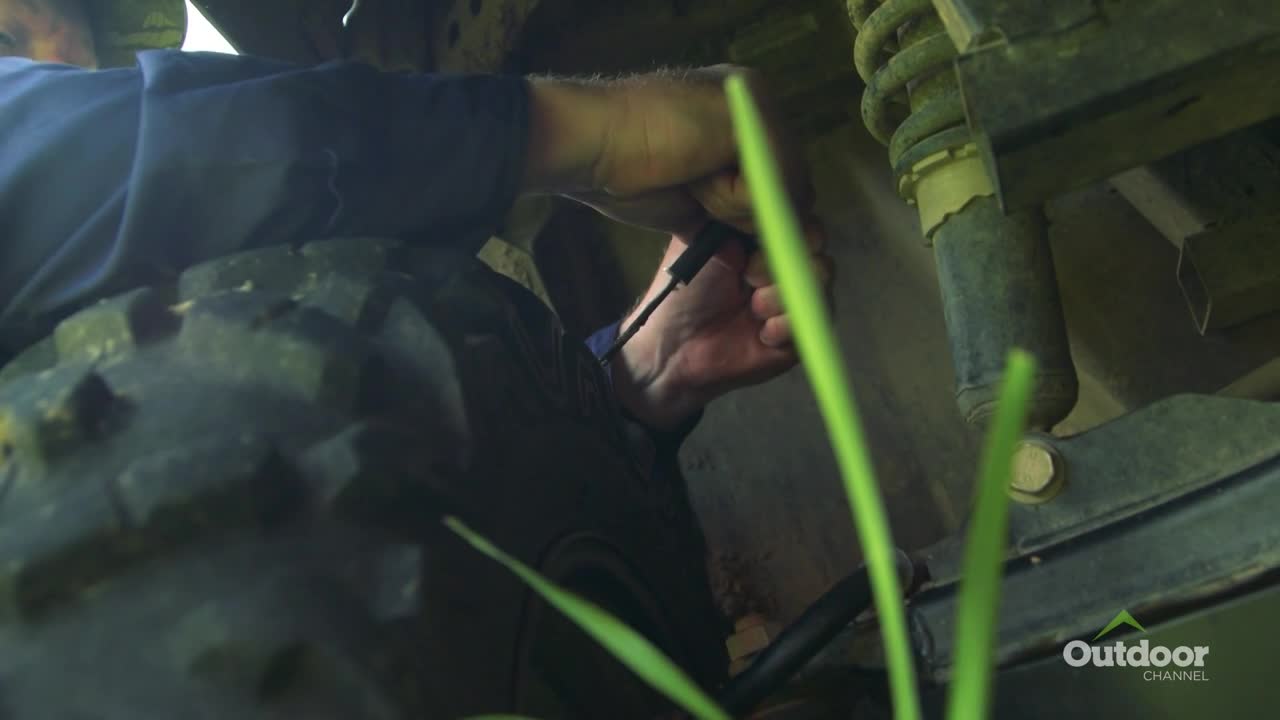A man's hand reaches up to work on the underside of a vehicle. He's holding a small black cable, likely plugging something in or unplugging it.
