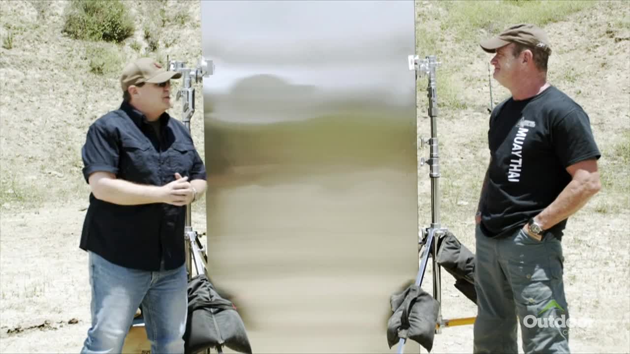 Two men stand on either side of a large, reflective panel, set up on tripods. The man on the left gestures with his hands while the other listens, hands in his pockets, with the Outdoor Channel logo visible.
