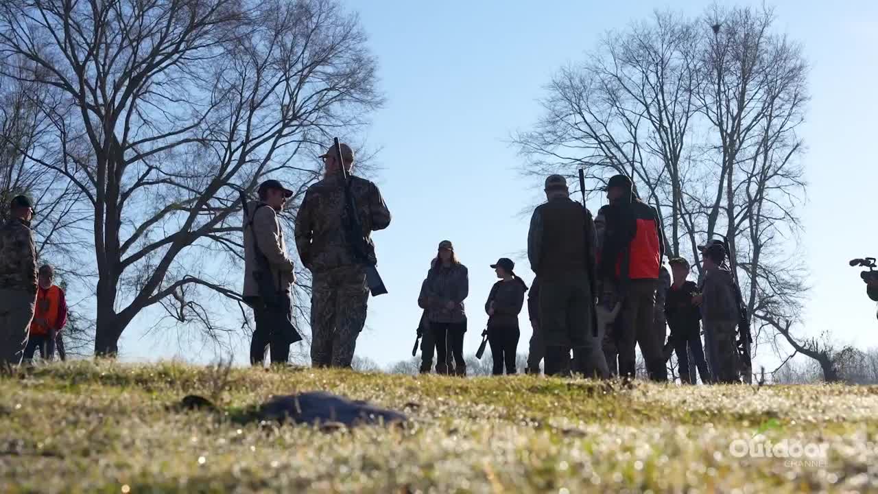 A group of people, some holding rifles, stand on a grassy hill under a bright, cloudless sky. The Outdoor Channel logo is visible, and the scene suggests a hunting or shooting activity.
