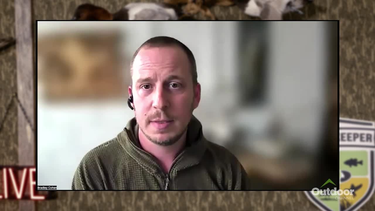 A man with a headset is speaking on a screen, framed by a backdrop of the Outdoor Channel logo and a "LIVE" sign. He's wearing a green jacket, and the background is blurred.
