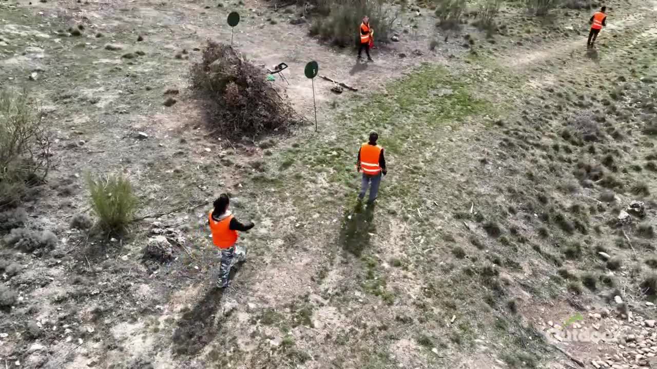 People in orange vests are moving across a grassy hillside, heading toward some targets. A few are already positioned near the shooting stations.
