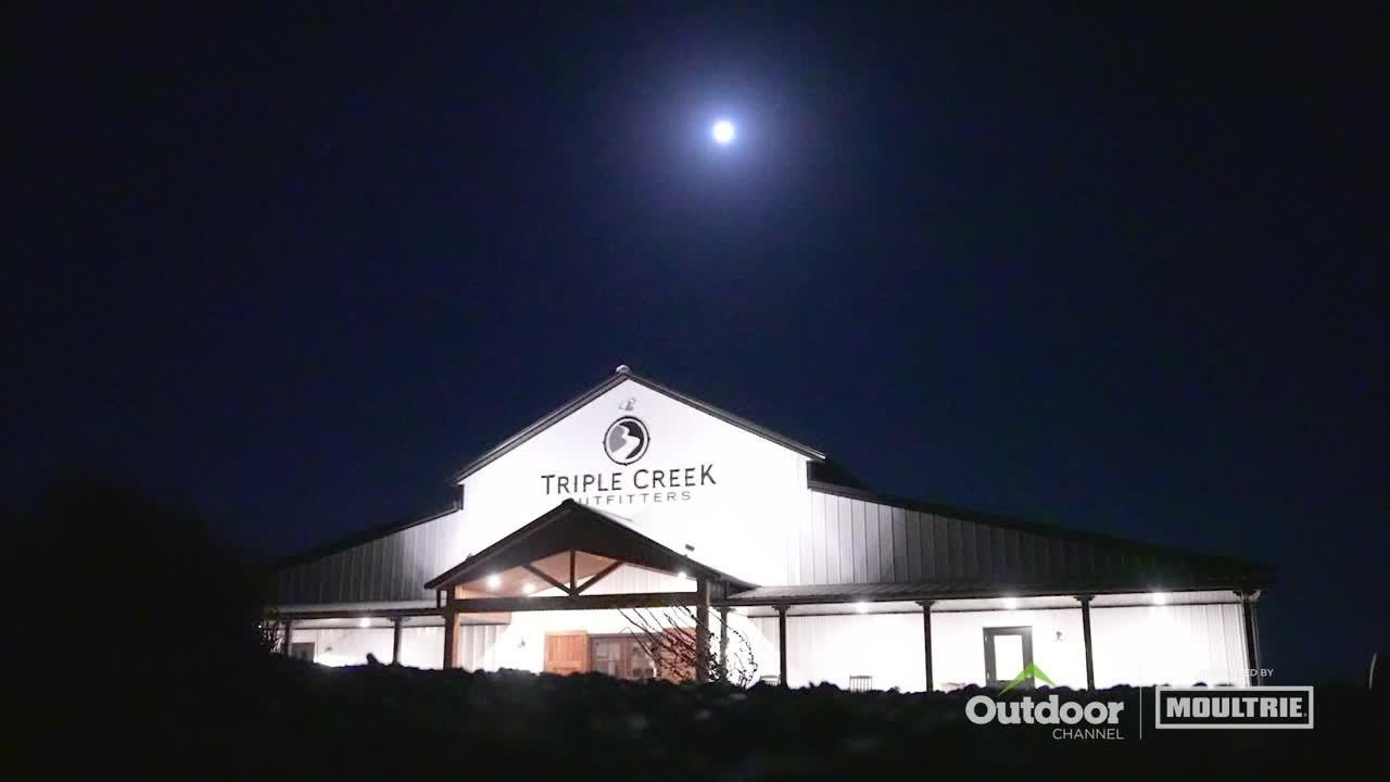 The moon hangs high above a large building lit up with the name "Triple Creek Outfitters." The Outdoor Channel logo is visible in the bottom right corner.
