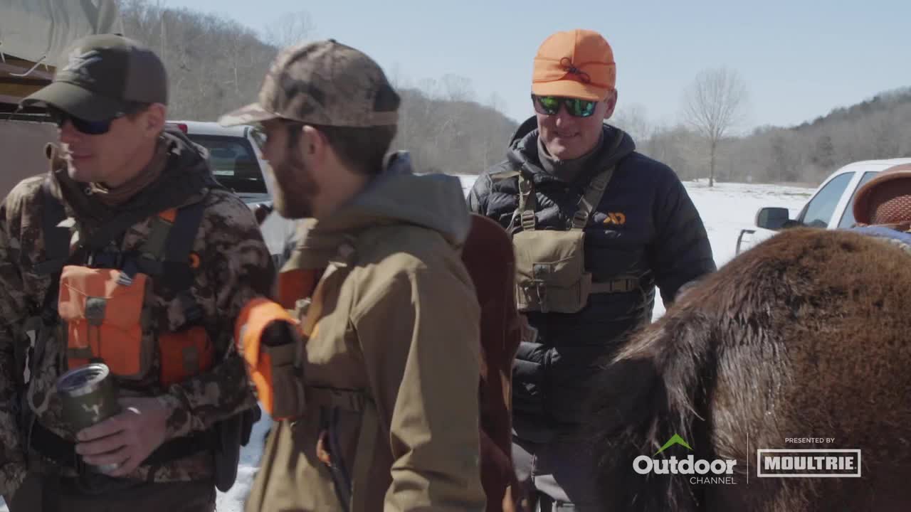 Three men in hunting gear stand near a large, brown bison, with snow on the ground behind them. The Outdoor Channel logo is visible at the bottom of the screen.
