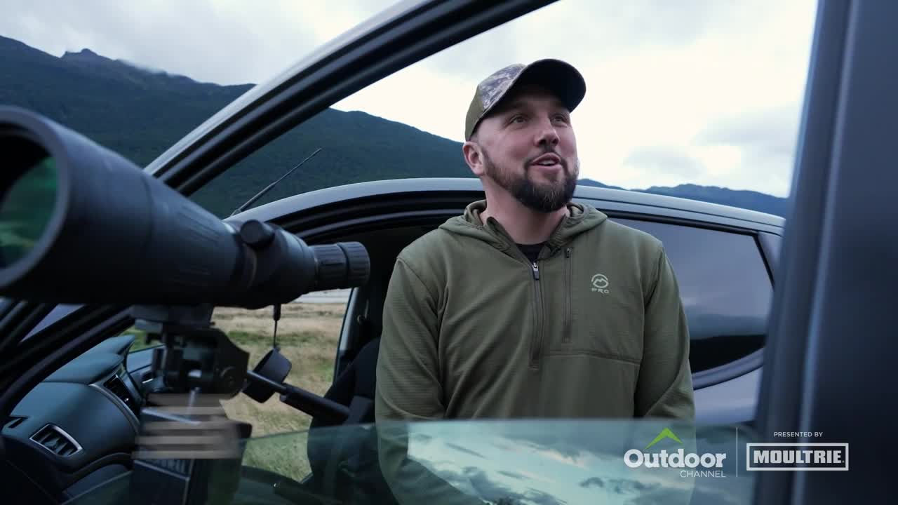 A man in a camouflage cap and green jacket stands beside a car, looking up and to his right. A large spotting scope is mounted inside the vehicle, pointed toward the mountains in the background.
