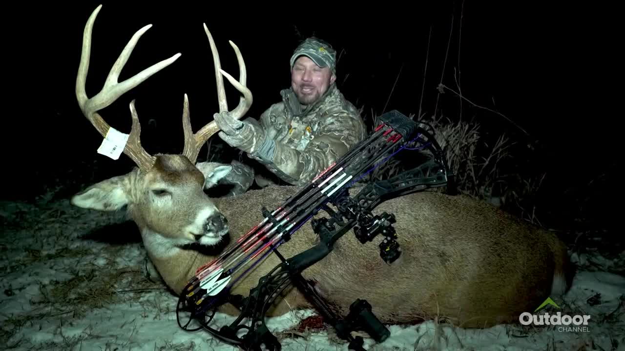 A man in camouflage kneels, smiling, over a large buck, its antlers towering.  A bow and arrows rest on the deer's side, and the Outdoor Channel logo is visible in the corner.
