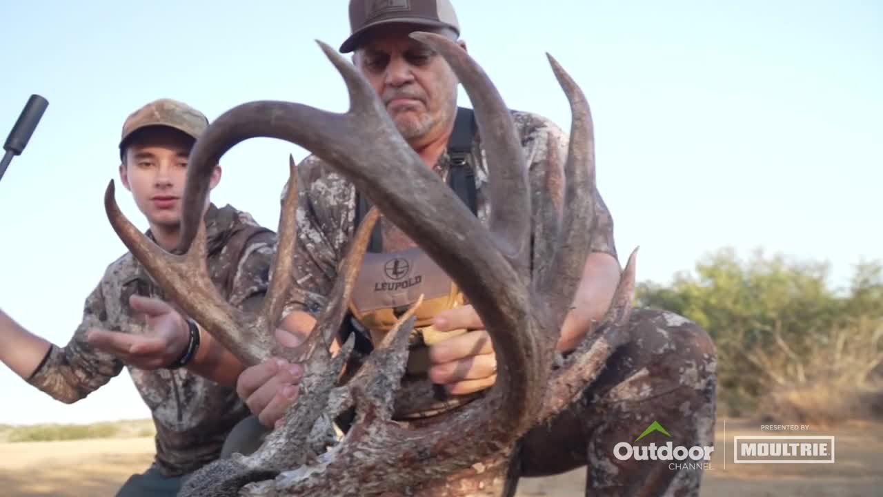 A man in camouflage holds a massive set of antlers, while his companion gestures towards them. The Outdoor Channel logo is visible in the corner.
