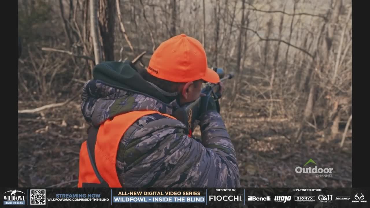 A hunter, clad in orange and camouflage, aims a rifle through the bare trees. The Outdoor Channel logo is visible, suggesting this scene is part of a hunting program.
