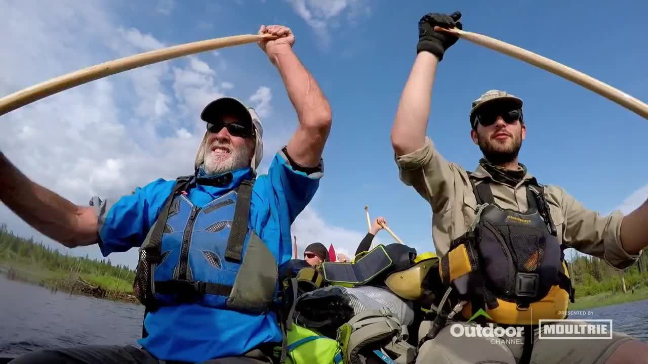 Two men in a canoe raise their paddles high, grinning at the camera. The Outdoor Channel logo is visible, suggesting this is part of a show about the outdoors.
