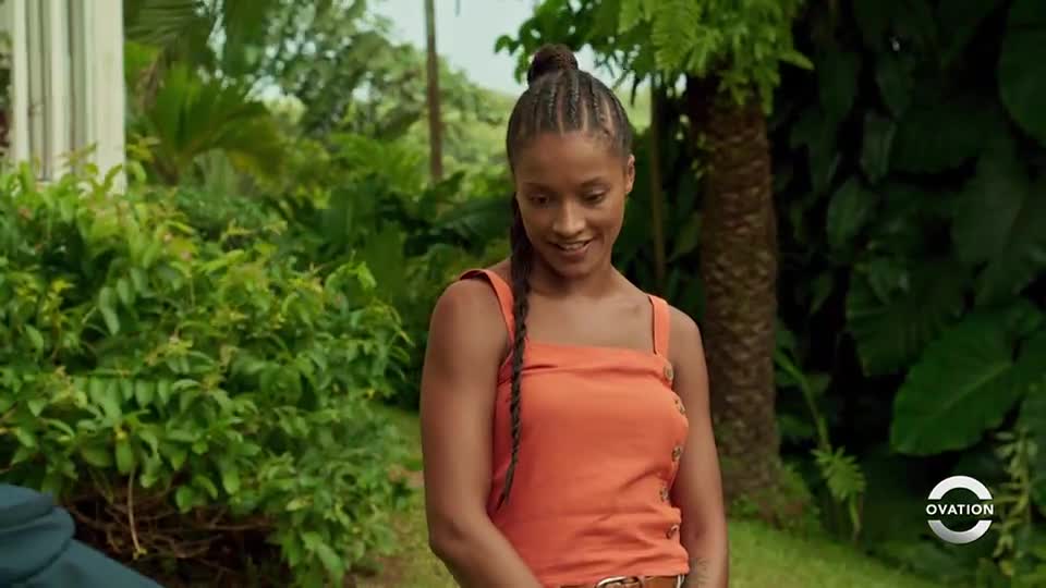 A young woman, wearing an orange top, smiles as she looks down. Lush greenery surrounds her in what appears to be a tropical setting, with the Ovation logo visible in the corner.
