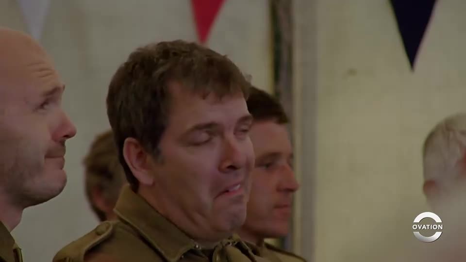 The man in the center has his eyes squeezed shut, mouth slightly open as if he's about to speak. He's surrounded by other men in uniform, the Ovation logo in the corner.
