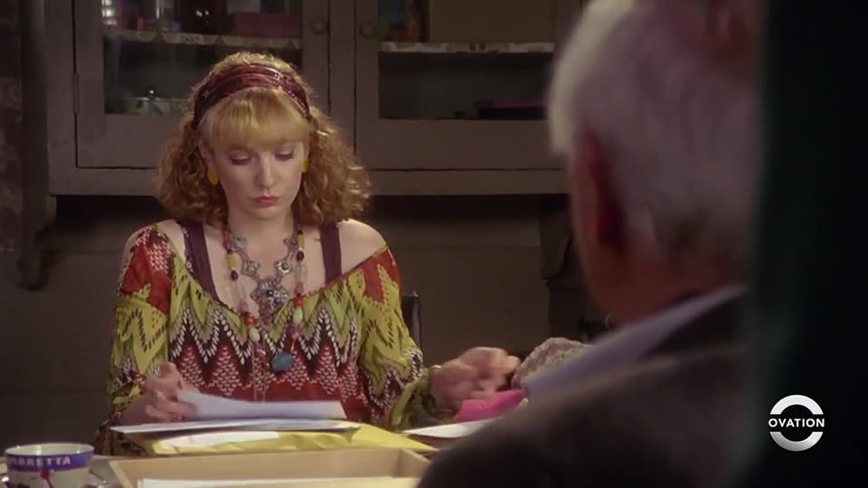 A woman with curly blonde hair examines papers at a table, while a man with white hair sits opposite her. The Ovation logo is in the corner.
