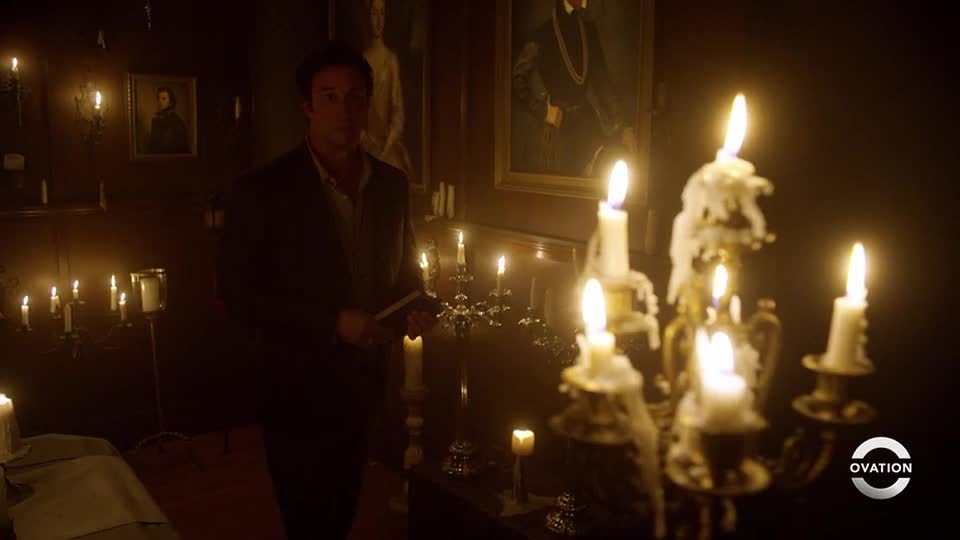 A man stands in a dimly lit room, holding a book while surrounded by lit candles. Thick wax drips down the sides of the candelabras, and the Ovation logo is visible in the corner.
