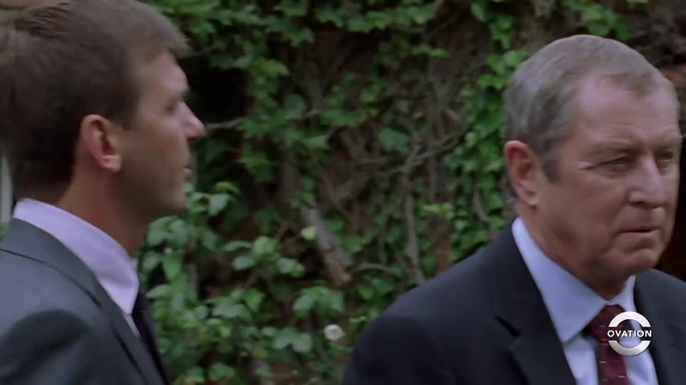 Two men in suits walk past a wall of green foliage. The older man's gaze is steady, while the other looks away.
