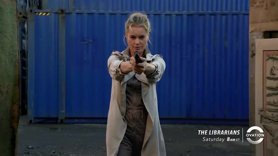 A woman in a trench coat holds a handgun, aiming directly at the viewer. Behind her, a large blue container provides a stark backdrop for the scene from *The Librarians* on Ovation.
