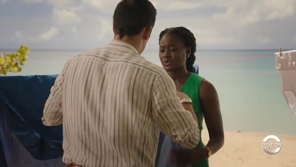 A man in a striped shirt is facing a woman in a green dress, their bodies close together. The woman has a concerned expression as she looks up at him.
