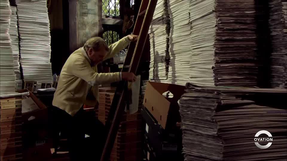 A man in a light jacket carefully climbs a wooden ladder, reaching towards a stack of papers. The room is filled with towering piles of documents, suggesting a vast archive.
