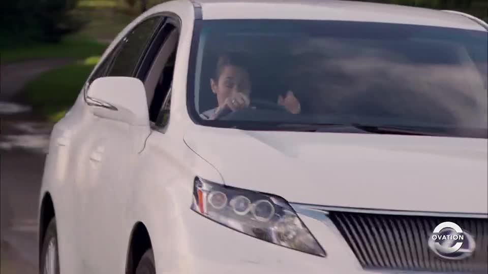 A white SUV is moving towards me, its headlights reflecting the sunlight. The driver, a woman, appears to be talking on the phone.

