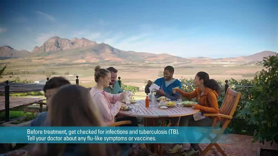 A group of people are seated around a table, eating and chatting outdoors. Mountains rise in the distance, and the sky is clear.
