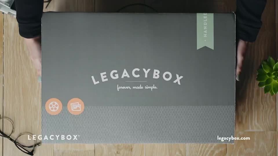 A person is holding a gray Legacybox, the top of which is facing the camera. The box is on a wooden surface, and the person's arms and hands are visible.
