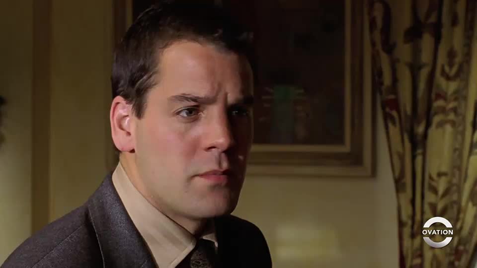 A man in a suit stares intently, his expression unreadable. The Ovation logo sits in the corner, suggesting a scene from a show or movie.

