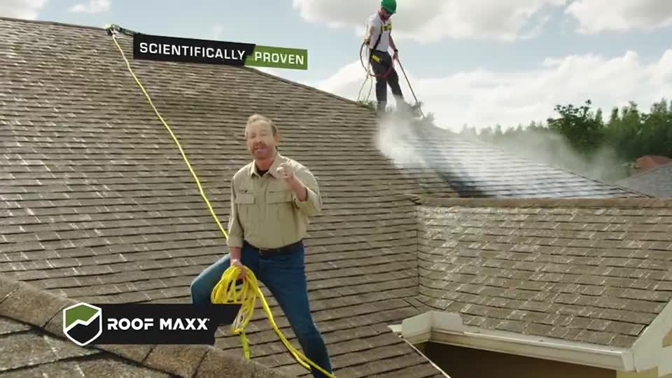 A man in jeans and a button-down shirt stands on a roof, holding a yellow cable, while another worker sprays the shingles with a high-pressure hose. Steam billows from the roof as the man gestures toward the camera.

