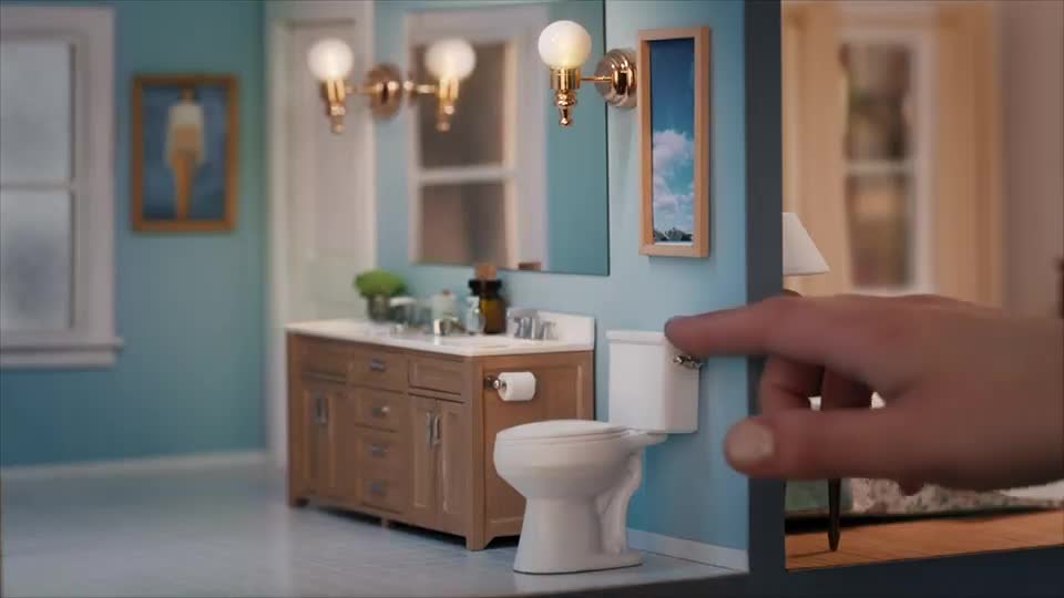 A finger presses the lever on a tiny toilet, part of a miniature bathroom. The scene is a detailed model, complete with a vanity and framed artwork.
