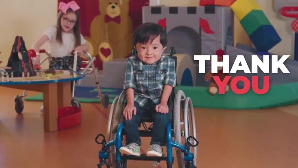 A young boy in a wheelchair smiles directly at me. Behind him, a girl with a pink bow plays with toys at a table.
