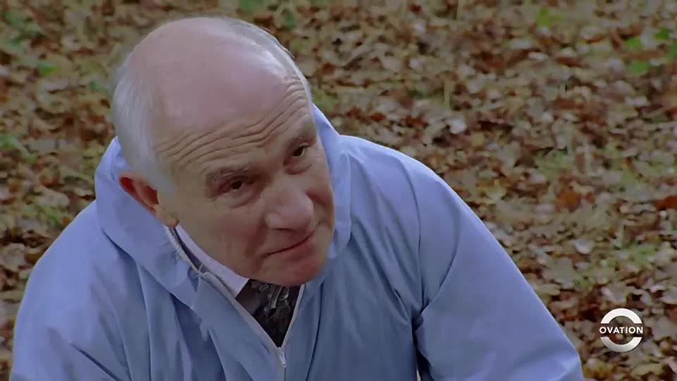 The man in the pale blue jacket looks directly at something just out of frame. He's in a wooded area, and the Ovation logo is visible in the corner.

