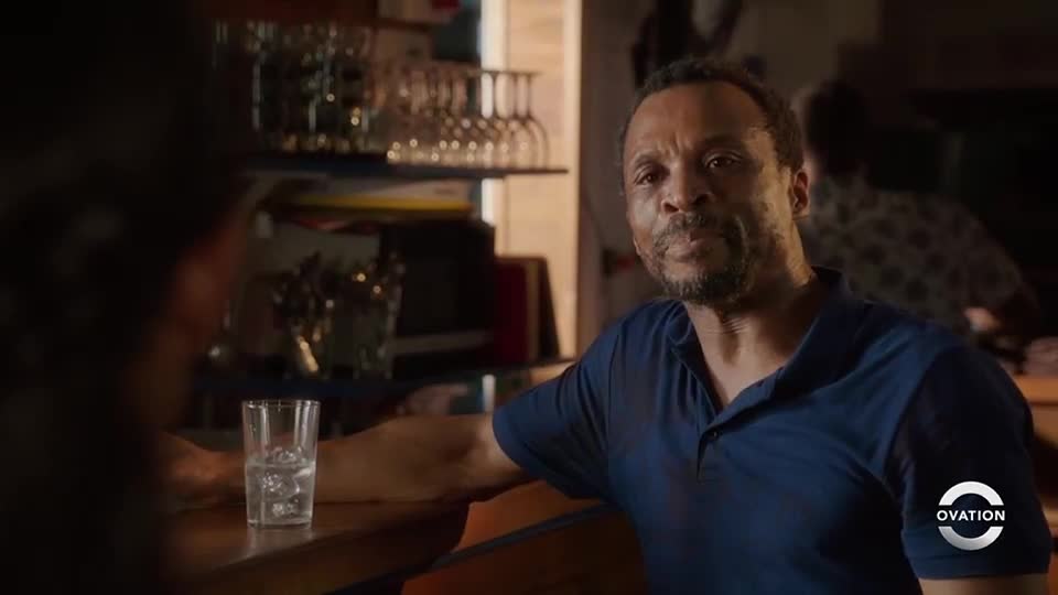 A man in a blue polo shirt leans on a bar, looking directly at someone out of frame. A glass of water sits on the counter beside him.

