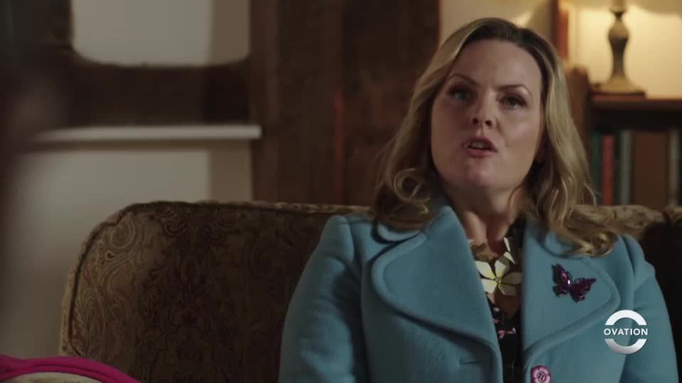 The woman, in a light blue coat, is speaking, her mouth open. A butterfly pin is affixed to her lapel, and the Ovation logo is visible.
