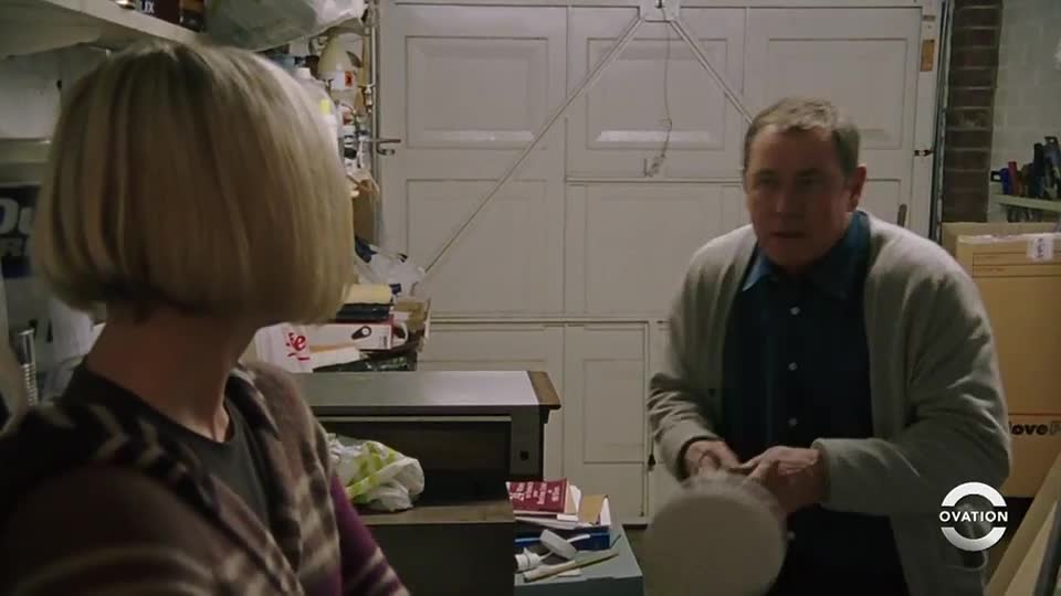 A man in a cardigan holds a round object, looking toward a woman with short blonde hair. They're in a cluttered garage, the Ovation logo visible in the corner.
