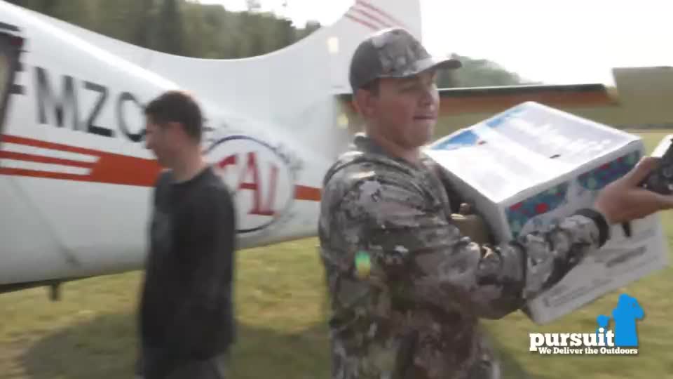 A man in camouflage carries a large white box towards the camera, walking past a small airplane. Another man in a black shirt walks near the plane.
