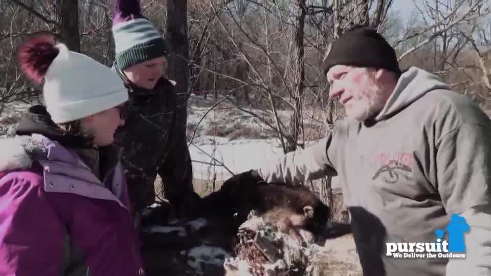 A man in a black hat gestures towards a dark-furred animal resting on a stump. Two children, bundled in winter coats, watch intently as he speaks.
