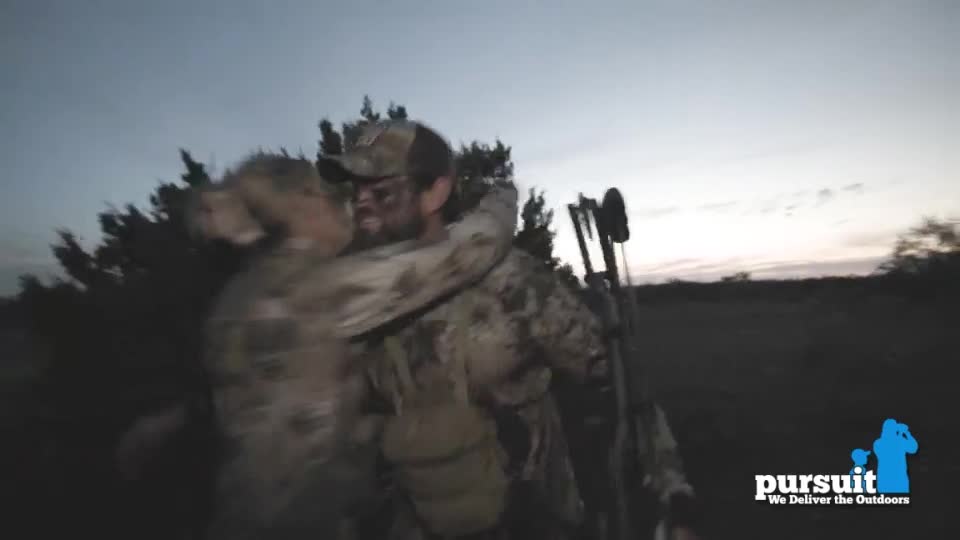 Two figures in camouflage embrace, their faces close together in the fading light. A bow and arrow are slung over the shoulder of one as they celebrate.
