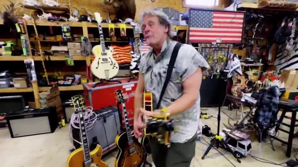 A man in a camo shirt strums a guitar, facing the camera in a cluttered workshop.  Behind him, an American flag hangs on the wall, and several guitars lean against amps.

