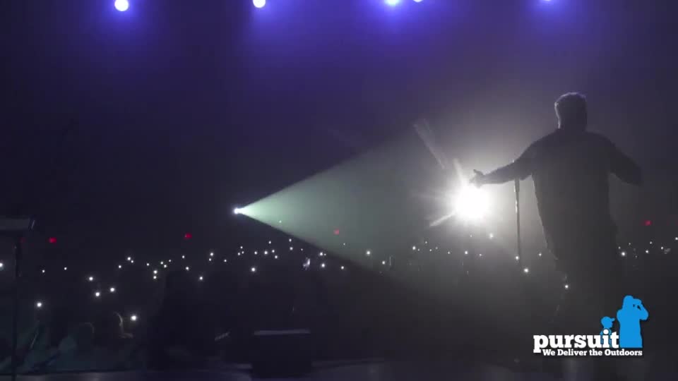 A spotlight shines on a performer, arms outstretched, as the crowd holds up their phones. The Pursuit Channel logo is visible in the corner.
