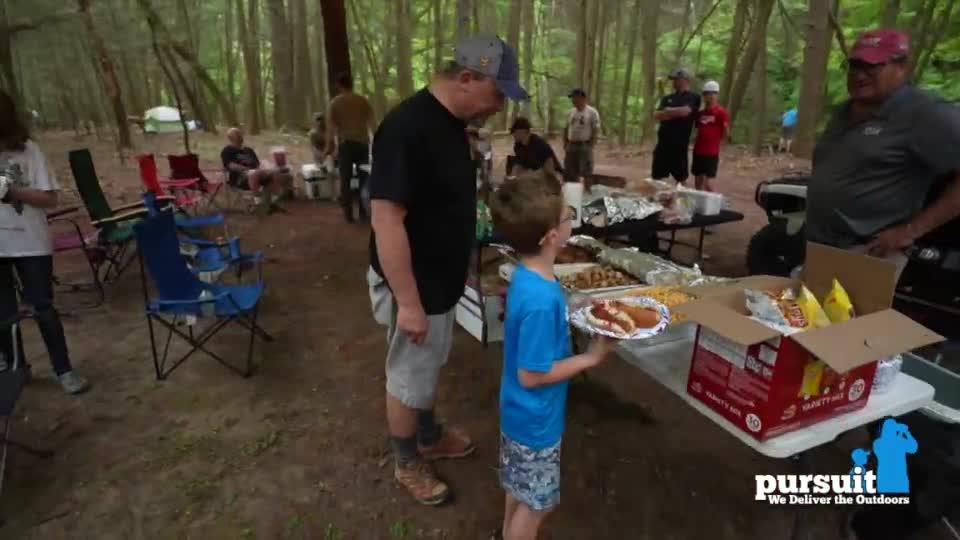 A young boy, holding a plate of food, stands near a table laden with prepared dishes. Men in baseball caps and casual clothes mill about, while others relax in folding chairs, all gathered in a wooded area.
