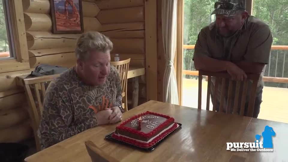 A man in camouflage blows out a single candle on a birthday cake. Another man, wearing a hat, watches from behind a chair.

