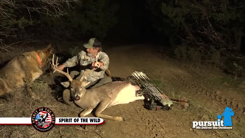 A man sits beside a fallen deer, his dog looking on, with a bow and arrows nearby. The scene appears to be filmed at night, likely for the Pursuit Channel, with the "Spirit of the Wild" logo visible.
