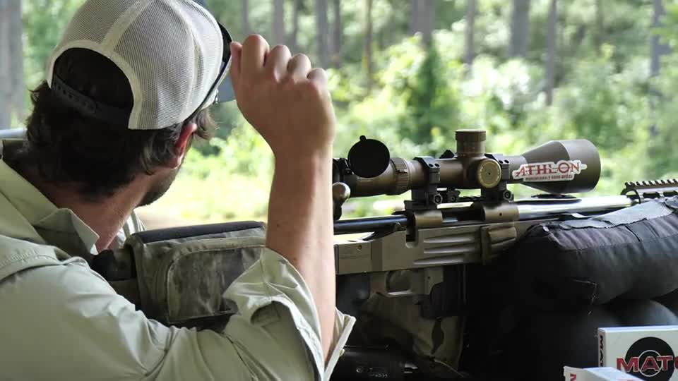 A man in a mesh-backed cap is adjusting his visor, peering through a scope mounted on a rifle. The rifle rests on a sandbag, ready for a shot.
