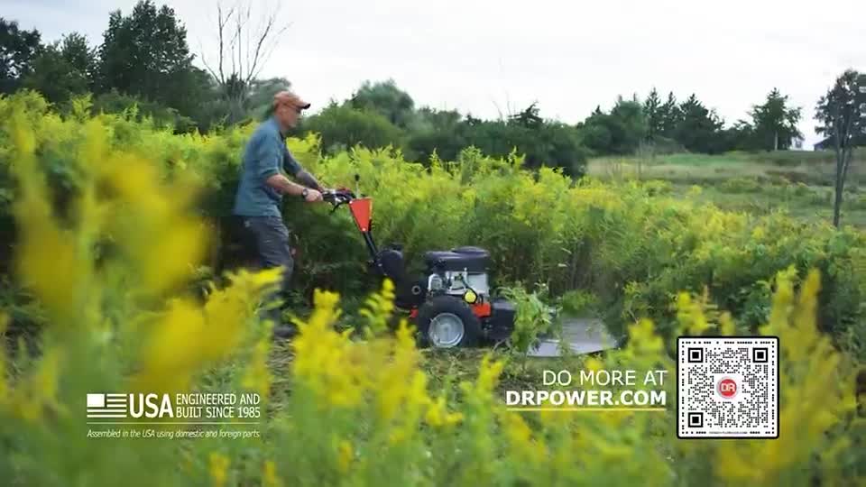 A man in a baseball cap pushes a small, orange mower through tall, yellow-flowered weeds. The machine cuts a path through the thick vegetation.
